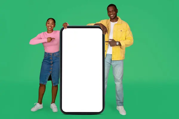 在线报价 积极可爱的美丽的千年黑人夫妇摆姿势 带着巨大的手机 白色空白屏幕 指向复制空间做广告 隔离在绿色工作室的背景下 — 图库照片