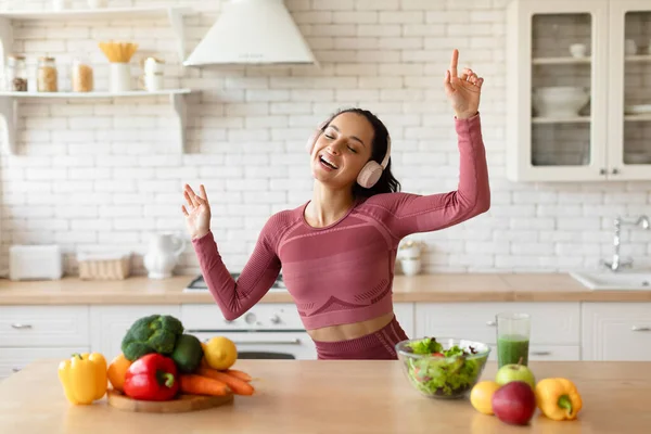 Healthy Weight Loss Lifestyle. Joyful Fit Woman Wearing Wireless Headphones, Dancing And Listening To Music Singing At Kitchen, Enjoying Slimming Routine Near Table With Healthy Vegetables