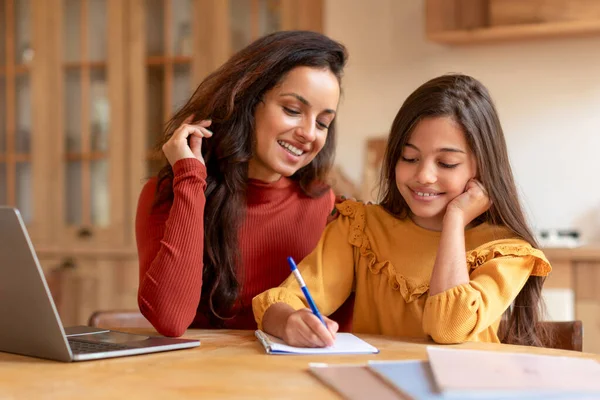 Homeschooling And E-Learning. Arabic Mom And Daughter Near Laptop Doing Kids Homework Together, Mother Or Teacher Helping Preteen Schoolgirl With School Studies, Schooler Taking Notes Indoor