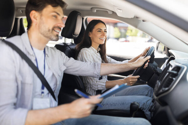 Driving school. Handsome man instructor examinating happy lady student, taking notes at chart while sitting by cheerful brunette woman driving auto, holding steering wheel, side view, copy space
