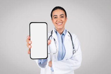 Gülümseyen Brezilyalı kadın doktor kameraya boş ekranlı akıllı telefon gösteriyor, gri arka planda tek başına duruyor, maket. Latin terapist kadın çevrimiçi tıbbi teklif öneriyor