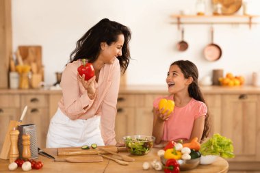 Neşeli bir Arap anne ve küçük kızı yemek pişiriyor ve gülüyor, taze sebze salatası hazırlarken, biber tutarken, mutfak masasının yanında dururken. Sağlıklı Yemek Tarifleri