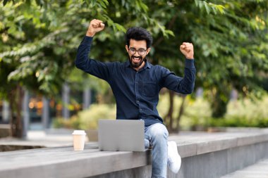 Heyecanlı, genç, doğulu girişimci bir adam sokakta uzaktan çalışıyor, bankta oturuyor, kahve içiyor, bilgisayarını kullanıyor, bilgisayar ekranına bakıyor ve el kaldırıyor, harika haber.
