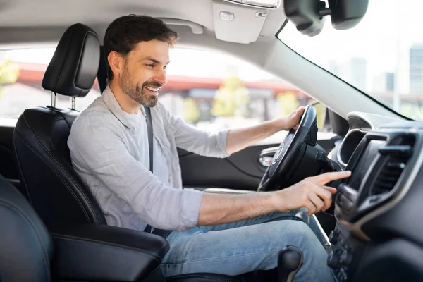 Happy cheerful handsome bearded young man wearing casual outfit driver sitting in front seat, holding auto steering wheel, turning on music on touch screen, driving his brand new nice car, copy space