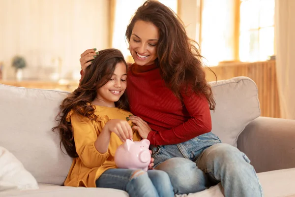 Family Finance Lessons. Arabic Mommy and Daughter Sharing Joy of Saving, Putting Money In Piggybank, Investing in a Kids Financial Future, Sitting On Couch In Modern Living Room Indoors