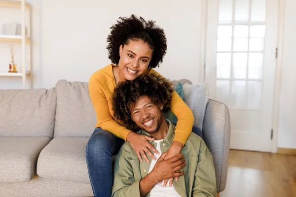 Happy African American Couple Sharing a Warm Embrace on the Sofa Indoor, Wife Hugging Husband From Back in Their Cozy Living Room, Smiling To Camera, Radiating Love and Happiness Together