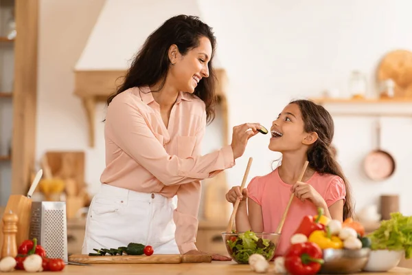 Cheerful Arabic Mommy And Her Daughter Kid Girl Cooking Together, Mom Feeding Preteen Child Giving To Taste Cucumber Slice, Having Fun While Preparing Family Dinner In Kitchen At Home Interior