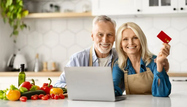 Online Shopping. Elderly Spouses Posing With Laptop And Credit Card In Kitchen, Smiling Senior Couple Making Payments In Internet Or Ordering Groceries Delivery, Enjoying E-Commerce, Closeup