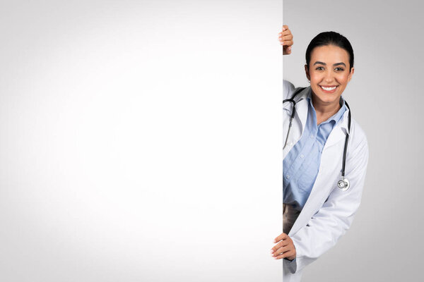 Medical ad. Happy latin female doctor peeking out of big blank advertisement board, cheerful brazilian therapist in uniform demonstrating empty space on white placard, panorama