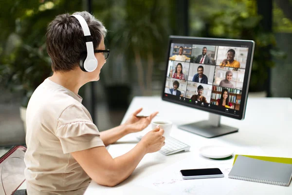 Middle aged woman remote employee have online meeting with international colleagues, working at coworking space, using computer pc and wireless headphones. Telecommunication, remote job