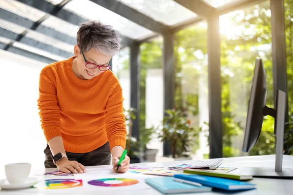 Creative mature woman interior designer working at home office, sun flare, copy space. Middle aged lady web-designer standing next to desk, working with color palette, making scetch, sun flare