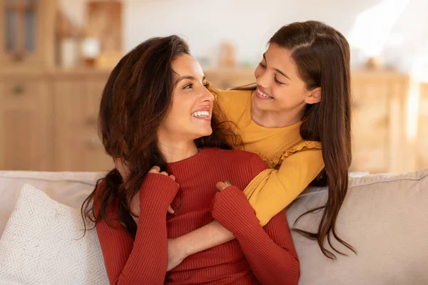 Mommy and Daughter Bond. Portrait of happy young arab mom and kid girl hugging, cute preteen daughter cuddling her mother from the back, sitting on couch at modern living room indoors