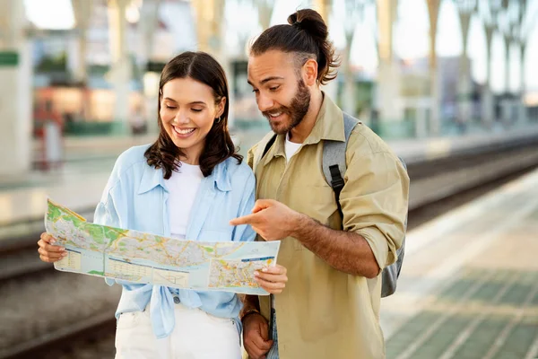 Cheerful young caucasian man and woman tourists in casual watching trip route on map on train station. Planning trip together and vacation, love, relationship and travel lifestyle