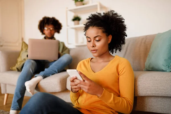 Internet Connected Family. Focused African American Woman Texting On Cell Phone While Husband Working On Laptop At Home, Young Serious Couple Engaging In Online Leisure. Technology. Selective Focus
