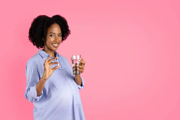 快乐千年怀孕的黑人女士与大腹部显示药丸胶囊和一杯水 隔离在粉红的背景下 工作室 维生素 矿物质 保健和医药建议 — 图库照片