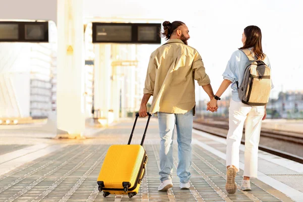 Happy millennial caucasian couple with suitcase hold hands, go to transport on train station, back, full length. Traveling together, active lifestyle, relationships and romance