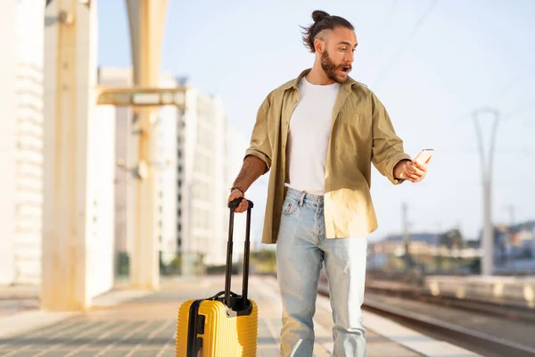 Shocked sad millennial caucasian man with suitcase looks on phone at train station. Hurry, problems in trip lifestyle, late for transport, human emotions, stress at vacation