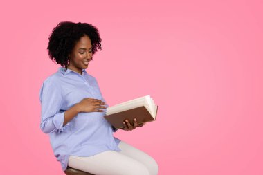 Milenyum siyahi hamile bayanın elleriyle karnına dokunması, kitap okuması, dinlenmesi, anneliğin tadını çıkarması, pembe arka planda, stüdyoda izole edilmesi. Beklenen çocuk, sağlık hizmetleri, hobiler, boş vakitlerde eğitim