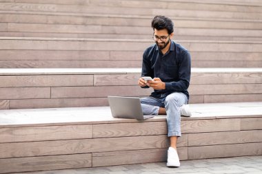 Yakışıklı Hintli Independent müteahhit dışarıda dizüstü bilgisayarda çalışıyor, telefon kullanıyor, şehir merkezindeki stadyumlarda oturuyor, e-postaları kontrol ediyor, kopyalama alanını kopyalıyor. Uzaktan çalışma, serbest çalışma