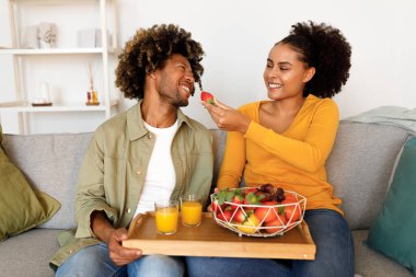 Romantik Atıştırma Zamanı. Taze meyve ve meyve suyunun tadını çıkaran siyah çifti seven, kocasını çilek ile besleyen, evdeki modern oturma odasındaki koltukta birlikte oturan,