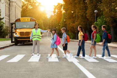 Road Traffic Safety. School Bus Assistant Lady Helping Kids To Cross Road By Crosswalk, African American Attendant Woman In Uniform Guiding Children, Group Of Pupils Walking On Pedestrian Crossing clipart