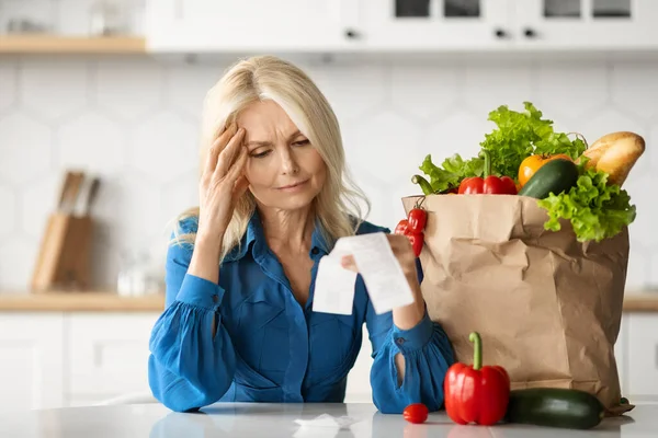 Grocery Expenses. Stressed Senior Lady Checking Bill After Food Shopping While Sitting At Table In Kitchen, Worry Mature Woman Concerned About Big Prices, Suffering Financial Crisis And Poverty