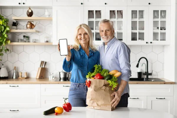 Smiling Senior Spouses Recommending Grocery Delivery App, Showing Blank Smartphone At Camera While Standing In Kitchen At Home, Happy Mature Couple Enjoying Shopping Application, Mockup