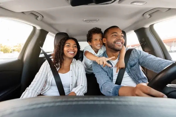 Beautiful cheerful loving african american family enjoying car trip. Cute preteen black boy embracing his father driving auto and pointing at road, happy parents and child spend weekend together