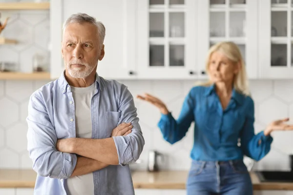 Selective focus on upset mature man with arms crossed on chest looking aside, having quarrel with his wife, kitchen interior. Crisis in marriage for seniors, divorce at older ages concept