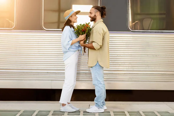Positive millennial caucasian couple meeting after trip, enjoy return, husband giving flowers to wife at train station. Travel end, relationships and romance, trip lifestyle