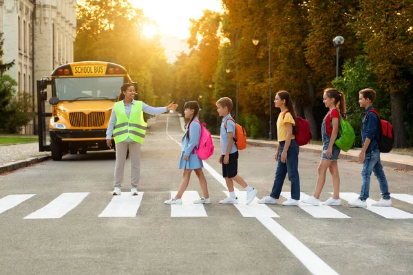 Road Traffic Safety. School Bus Assistant Lady Helping Kids To Cross Road By Crosswalk, African American Attendant Woman In Uniform Guiding Children, Group Of Pupils Walking On Pedestrian Crossing