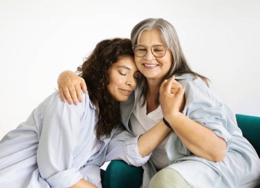 Beyaz tenli yaşlı bir anne yetişkin bir kadına sarılıyor, beyaz duvar arkasında oturma odasında sevgi ve desteğin tadını çıkarıyor. Evde ilişkiler, bakım, anne-kız ilişkileri, reklam ve sunum