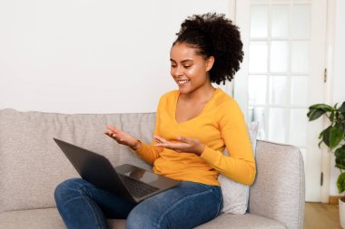 Çevrimiçi İletişim. Laptop bilgisayarlı neşeli Afrikalı Amerikalı kadın video görüşmesi yapıyor, koltukta oturuyor, çalışıyor ya da evdeki Modern Oturma Odasında uzaktan öğreniyor. Serbest ve Teknoloji