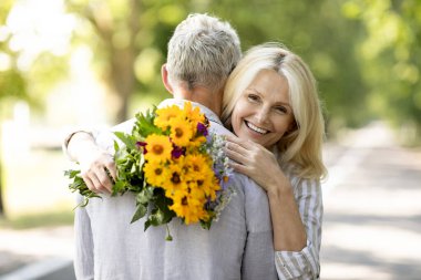 Yaşlılıkta romantizm. Mutlu Olgun Kadın Çiçekleri Tutuyor ve Dışarıda Birlikte Beklerken Kocasına Kucaklıyor, Romantik Üst Düzey Eşler Dışarıda Çıkıyor, Yıldönümlerini Kutluyor