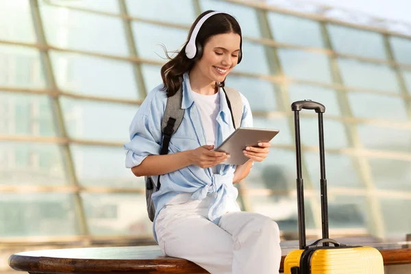 Positive millennial caucasian lady with suitcase in headphones, chatting on tablet at airport, train station. Travel blog, online booking, trip lifestyle, human emotions and music