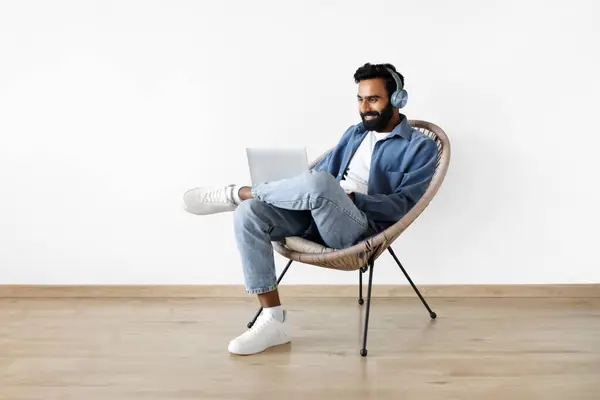 Online Work, Freelance. Indian Bearded Guy With Laptop Sitting Wearing Headphones, Video Calling And Working Remotely, Sitting In Comfortable Armchair At Home Near White Wall, Full Length