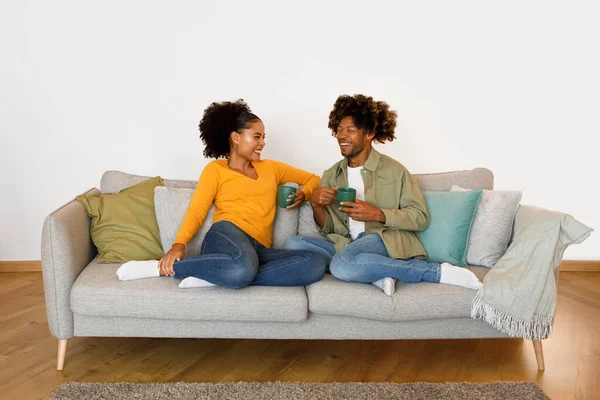 Coffee Break Together. Smiling Married Black Couple Cuddled Up on Couch, Sipping Coffee and Enjoying Conversations And Flirt on Relaxing Weekend at Home Interior. Cozy Romantic Moments. Front View