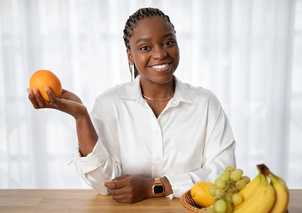 快乐的年轻黑人女士手握橙子 对着相机微笑 坐在餐桌旁 盘中放着各种水果 漂亮的女人享受着健康的生活方式和富含维生素的饮食 复制空间 — 图库照片