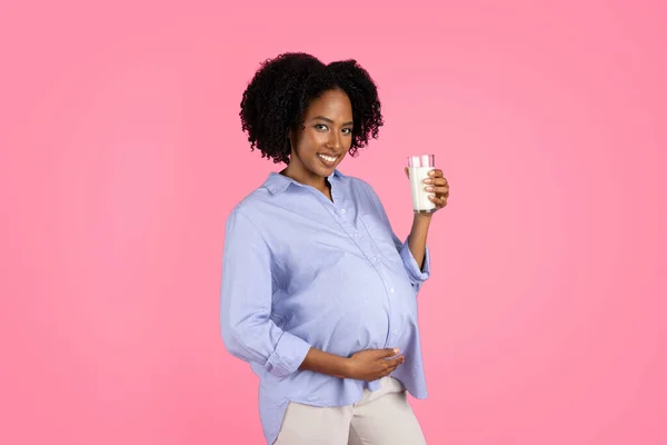 Cheerful millennial black lady with big belly drinking glass of milk, isolated on pink background, studio. Calcium and vitamins, health care of unborn child, diet and lifestyle