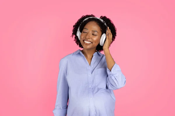 Cheerful millennial black lady with big belly in wireless headphones listening to music, enjoy spare time, isolated on pink background, studio. Lifestyle, device for rest and relax alone