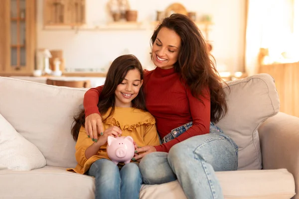 Kid Personal Savings. Happy Arabic Mom And Daughter Filling Piggybank with Coins on Couch At Home Interior, Mother Hugging Preteen Child Teaching Her Financial Literacy Indoors