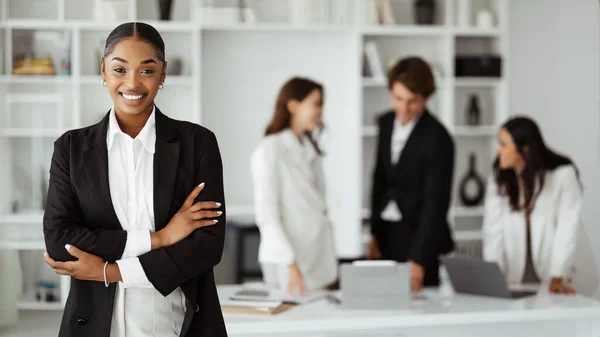 Happy young black businesswoman posing with folded arms and smiling, standing in conference room in office interior during meeting with colleagues