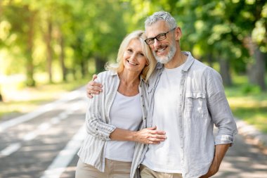 Happy Senior Couple Embracing And Smiling To Camera While Posing Together Outdoors, Cheerful Mature Spouses In Casual Clothes Standing In Sunny Park, Enjoying Retirement Lifestyle, Copy Space clipart