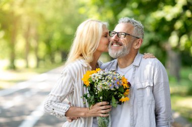 Güzel, olgun bir kadın çiçekleri tutuyor ve parkta yürürken kocasını öpüyor, kıdemli eşlerini dışarıda romantik bir şekilde buluşuyor, dışarıda yürürken kaynaşıyor, boşluğu kopyalıyor.