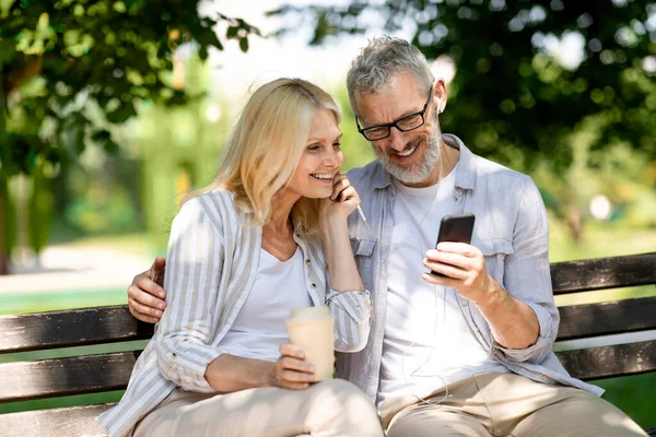 Happy Senior Couple With Smartphone Relaxing Together On Bench In Park, Cheerful Mature Man And Woman Listening Music On Mobile Phone, Sharing Earphones And Drinking Takeaway Coffee, Free Space