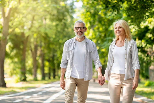 stock image Happy mature man and woman holding hands and walking on path in park together, smiling romantic senior couple spending time outside, smiling and laughing, enjoying outdoor date, copy space