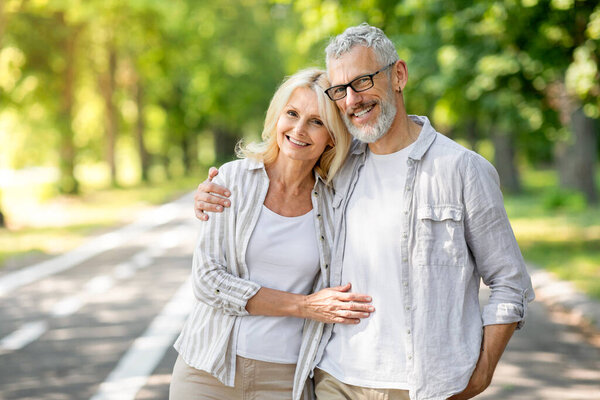 Happy Senior Couple Embracing And Smiling To Camera While Posing Together Outdoors, Cheerful Mature Spouses In Casual Clothes Standing In Sunny Park, Enjoying Retirement Lifestyle, Copy Space