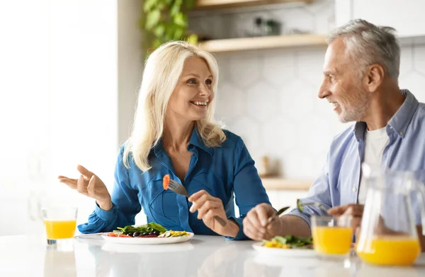 Romantic senior couple eating tasty breakfast together in kitchen at home, happy elderly spouses enjoying delicious food and drinking orange juice, sitting at table, chatting and laughing