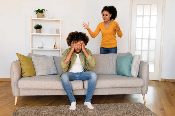 Unhappy black husband covers his eyes and ears in frustration as angry wife shouts at him during heated quarrel at home. Young african american couple facing marital crisis issue. Selective focus