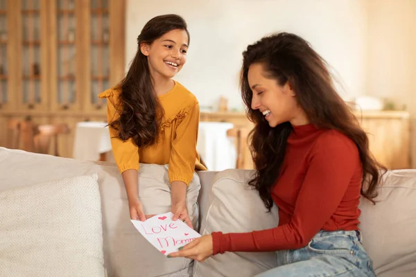 Mothers Day Surprise. Cheerful Arabic Preteen Kid Daughter Congratulating Mom On Holiday Or Birthday, Giving Handmade Greeting Card Painting To Her, Celebrating In Modern Living Room At Home
